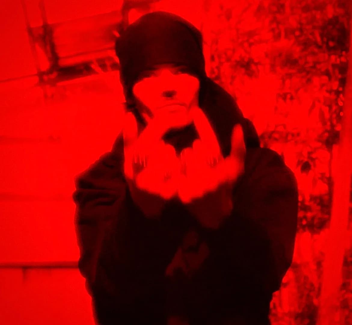 Red-tinted photograph of a hooded figure gesturing with both hands toward the camera