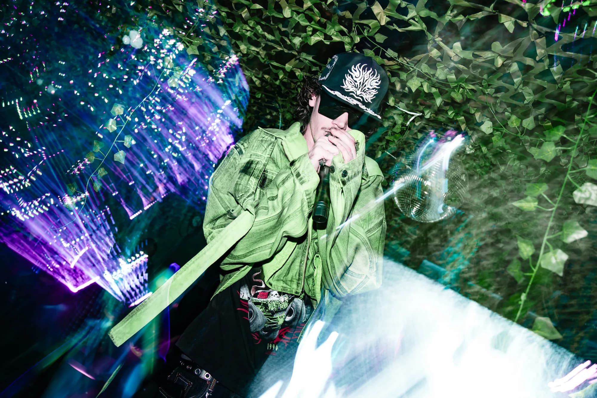 Performing in a green jacket and black cap under camouflage netting and stage lights