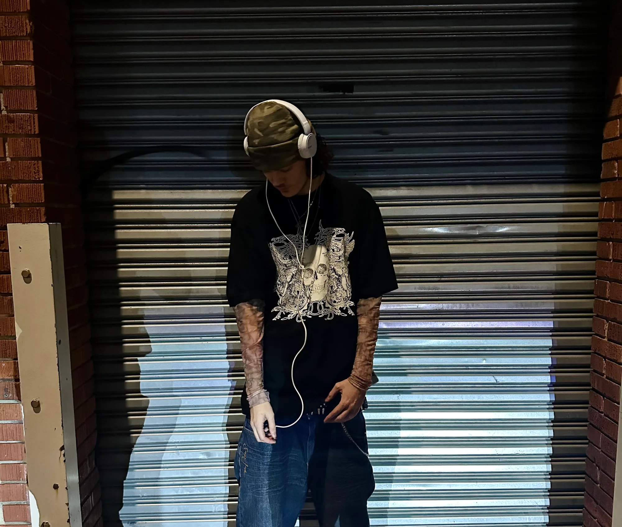 Leaning against a metal roller shutter, wearing headphones and a camouflage cap