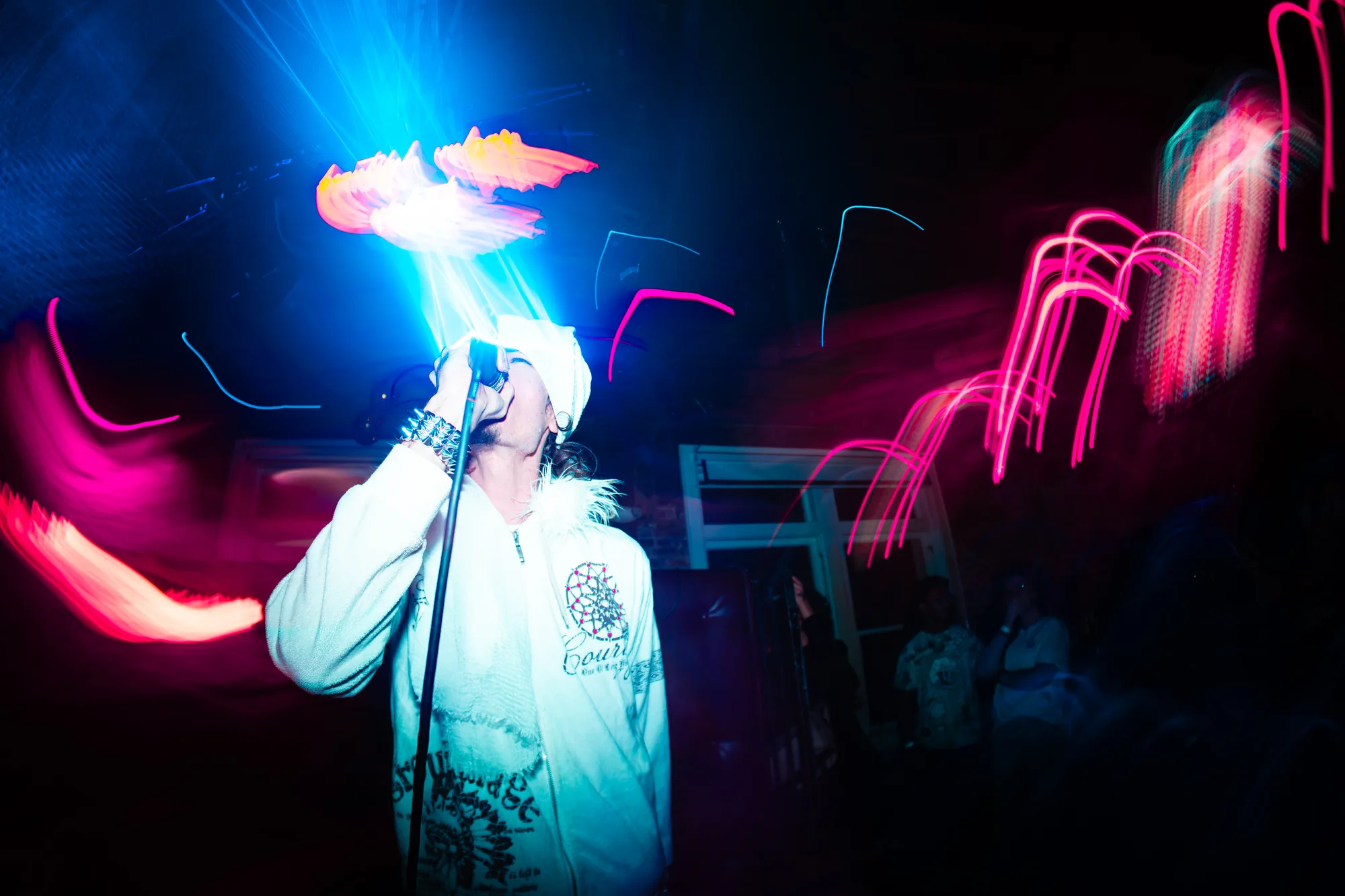Singing into a microphone in a white hoodie amid streaks of colored light