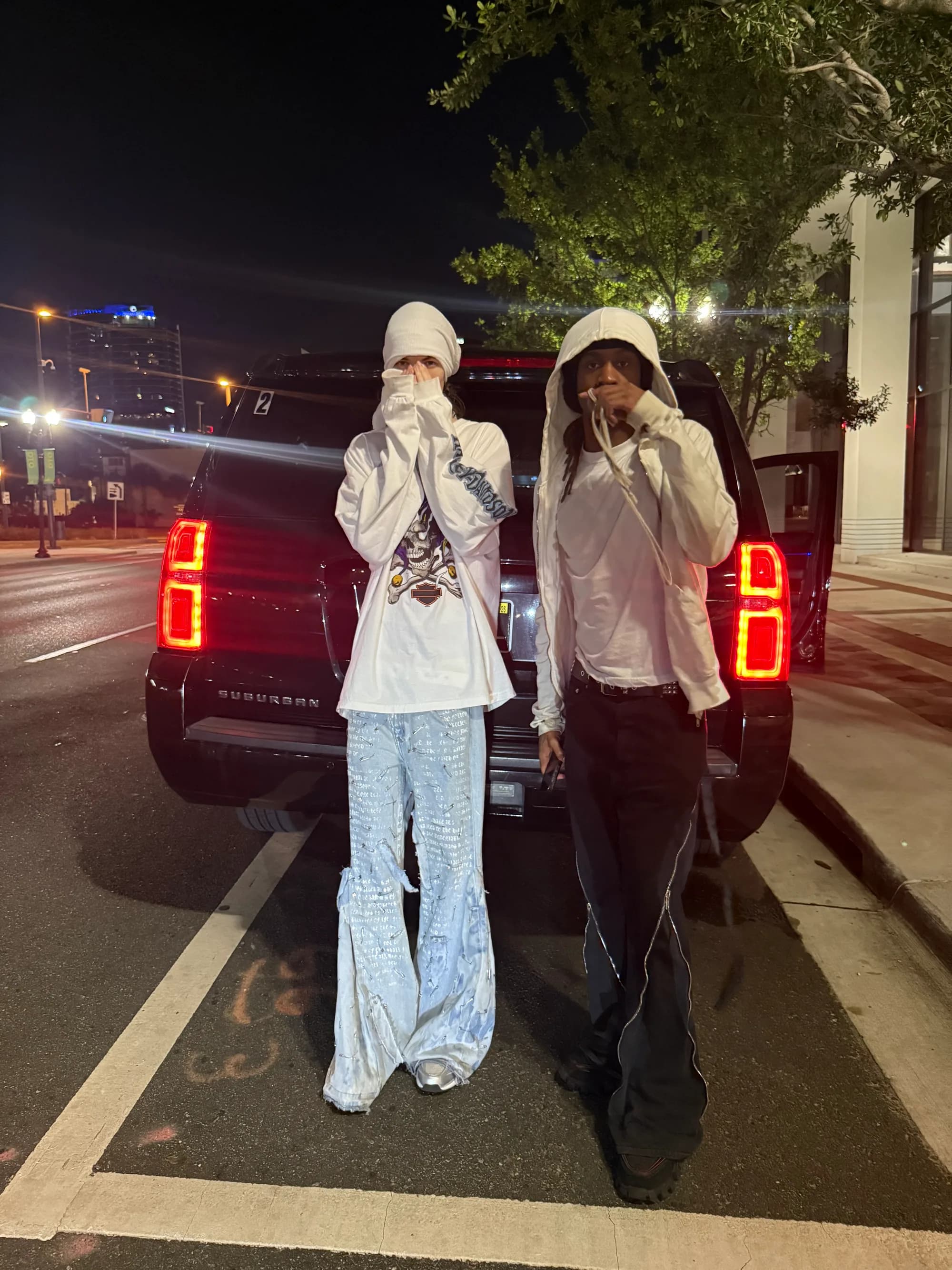 Two figures in white hoods standing behind a black SUV at night