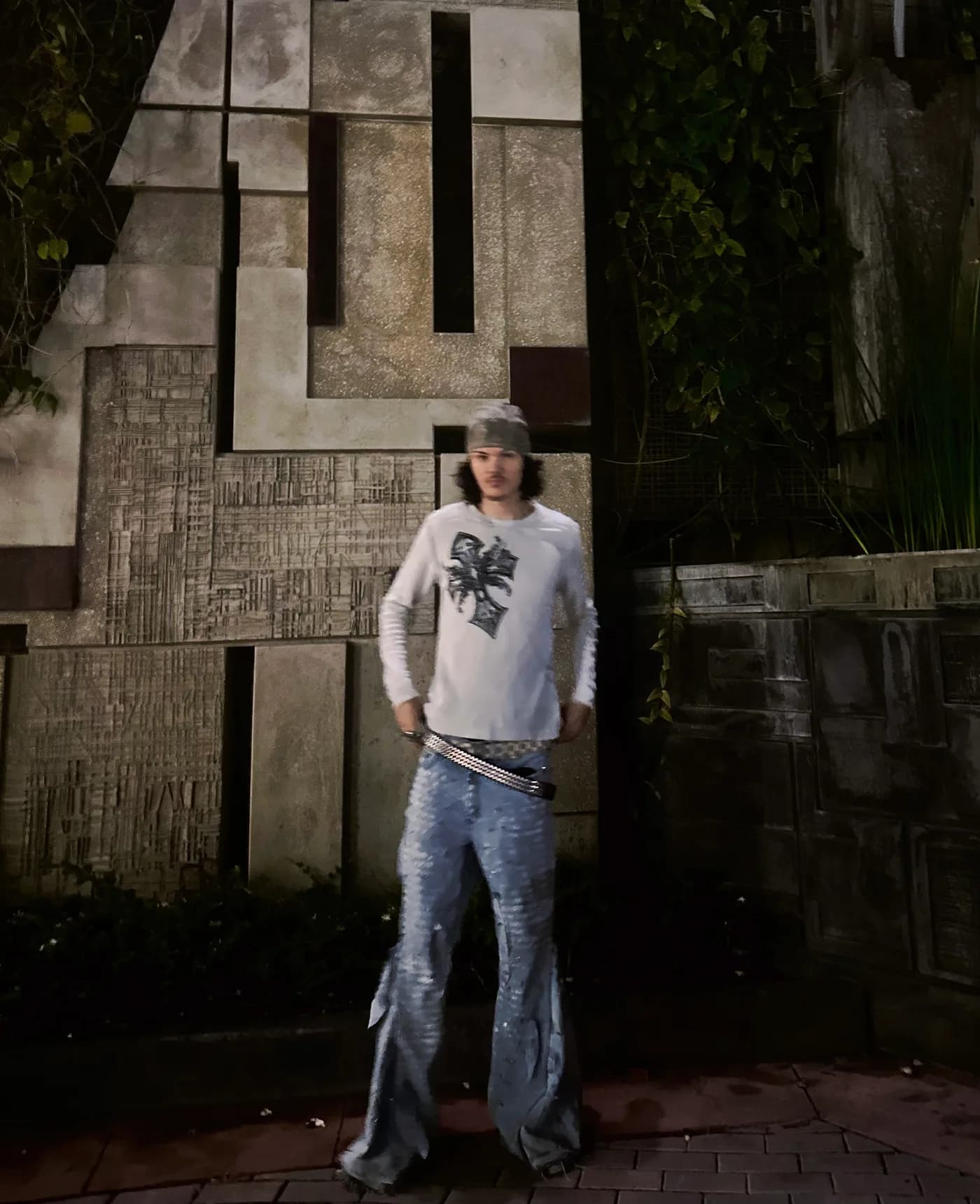 Standing before a carved concrete wall in a white long-sleeve shirt and patterned jeans